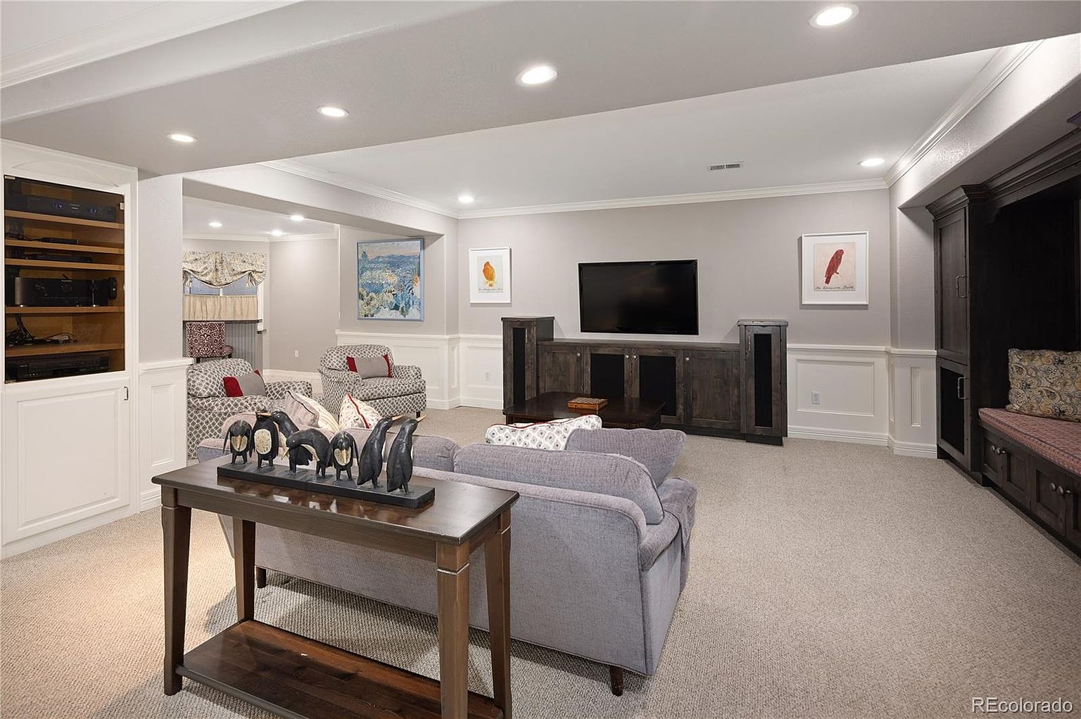 This spacious, finished basement features a comfortable living area with a plush grey sofa, patterned armchairs, and a large media console centered around a wall-mounted television. The room is accented by built-in cabinetry, wainscoting, and decorative artwork, creating a cozy and inviting atmosphere. The perspective is from behind a console table, looking into the main seating area, highlighting the open layout and neutral color palette.