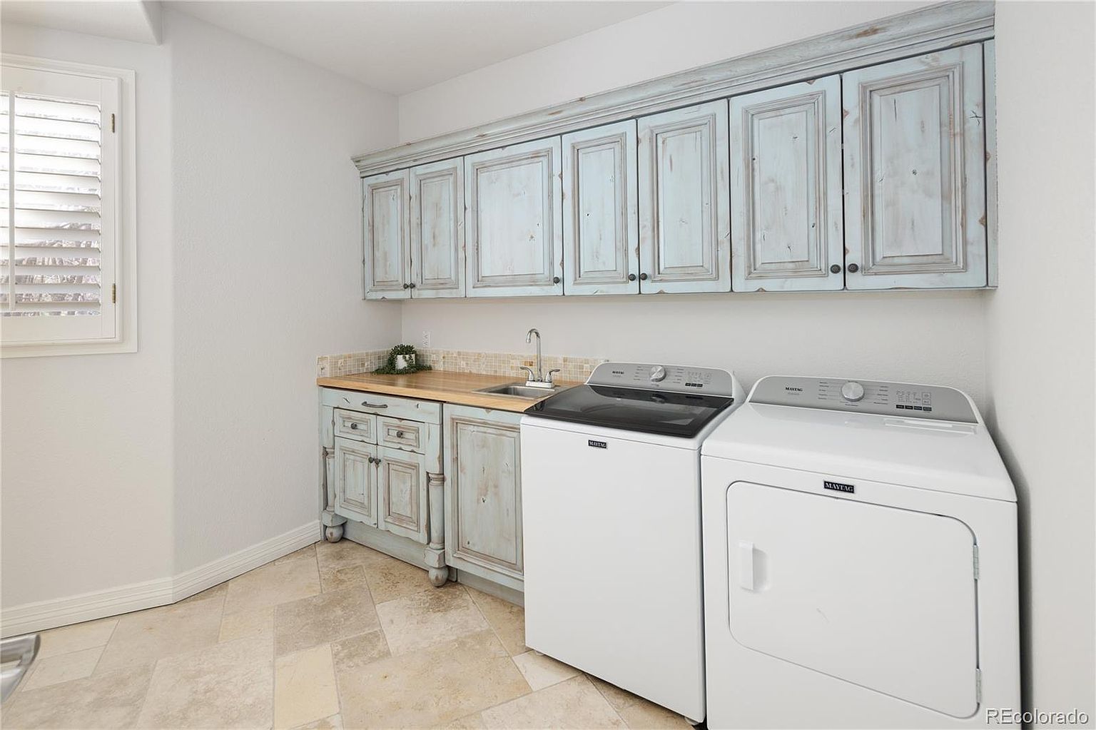 This laundry room features a rustic, distressed blue-grey cabinetry set that provides ample storage above a wooden countertop and a small utility sink. The space is equipped with a white Maytag washer and dryer set, set against a neutral wall and tiled flooring. The room is bright and functional, offering a clean and organized space for household chores.