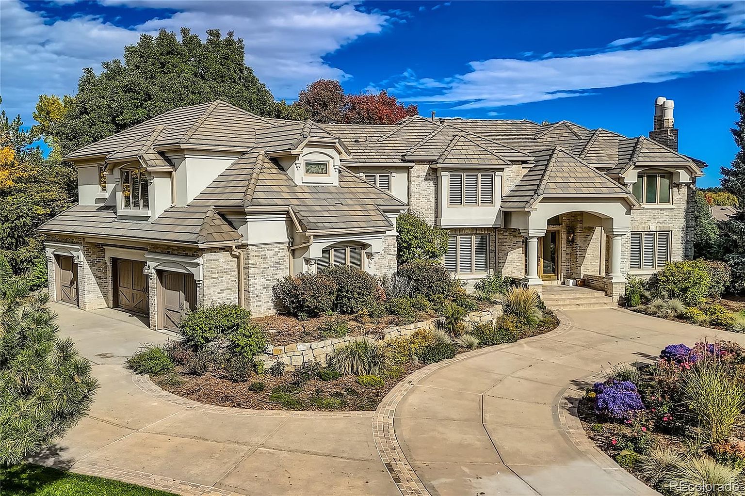 This grand, multi-story luxury residence features a sophisticated blend of light brick and stucco siding, complemented by a complex, multi-gabled roofline. A sweeping concrete driveway leads to a three-car garage on the left and a welcoming, covered entryway on the right, all set against a backdrop of mature trees and manicured landscaping. The perspective is a slightly elevated eye-level shot, capturing the home's impressive curb appeal and architectural depth on a bright, sunny day.