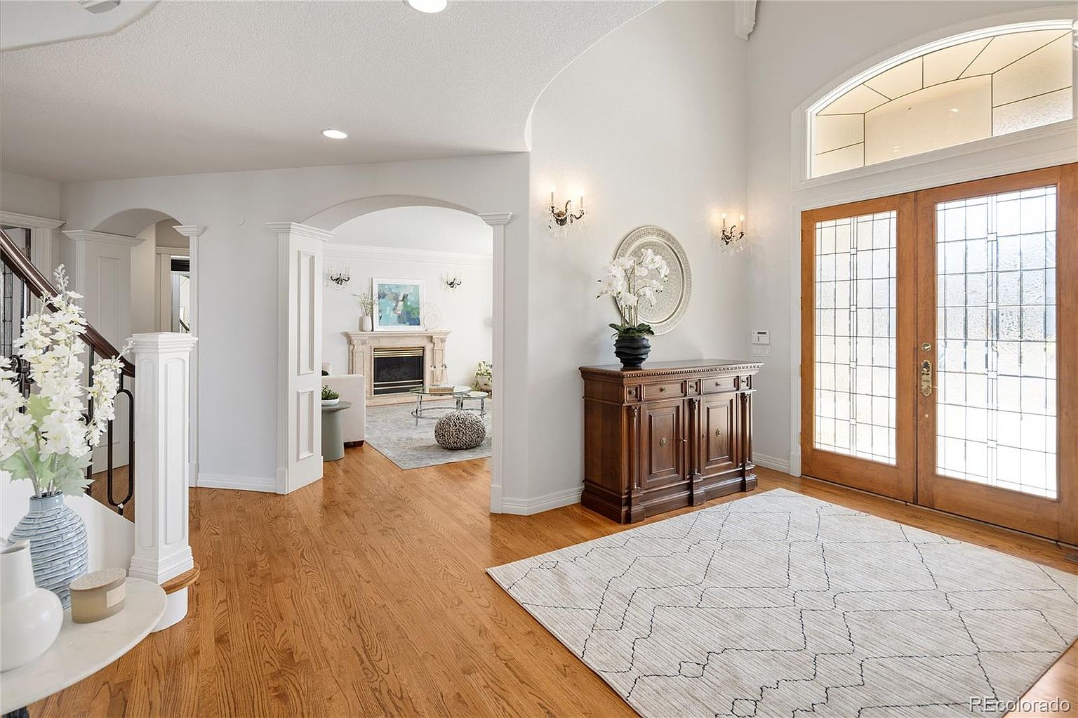 This elegant entryway features warm hardwood flooring, a grand staircase with white balusters, and a double-door entrance with decorative glass panels. The space flows seamlessly into a bright, open living area with a fireplace, creating an inviting and sophisticated atmosphere. The neutral color palette and high ceilings enhance the sense of openness and luxury throughout the home.
