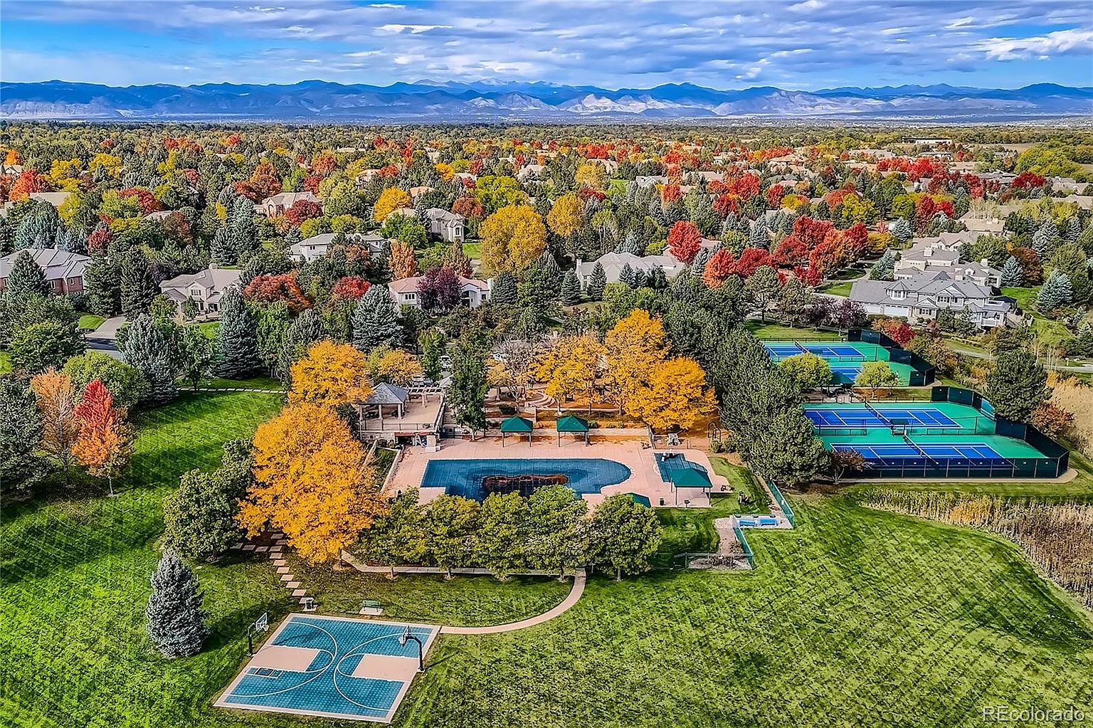 This high-angle aerial view captures a vibrant community recreational area nestled within a lush, tree-lined neighborhood during autumn. The facility features a large swimming pool, multiple tennis courts, and a basketball court, all surrounded by expansive green lawns and colorful foliage. In the background, a scenic mountain range provides a stunning backdrop to this well-maintained residential amenity space.