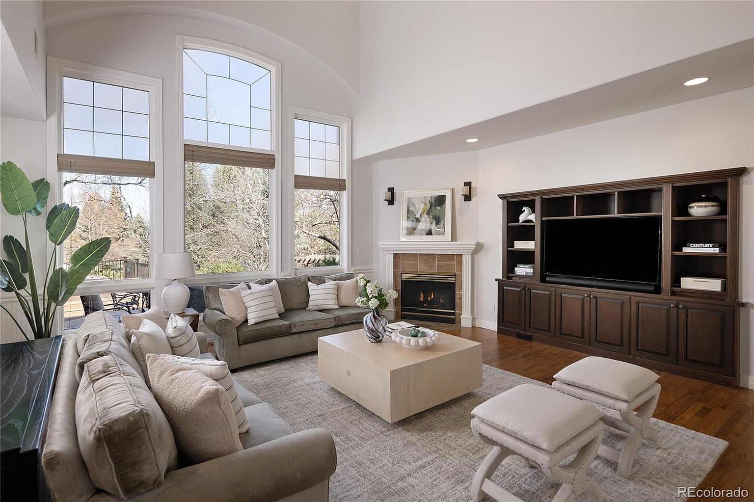 This spacious, light-filled living room features a soaring vaulted ceiling and large windows that provide an airy, open atmosphere. The room is anchored by a cozy fireplace with a stone surround and a built-in dark wood entertainment center, complemented by comfortable neutral-toned seating and a large, light-colored coffee table. The overall aesthetic is warm and inviting, blending traditional architectural elements with contemporary furnishings.