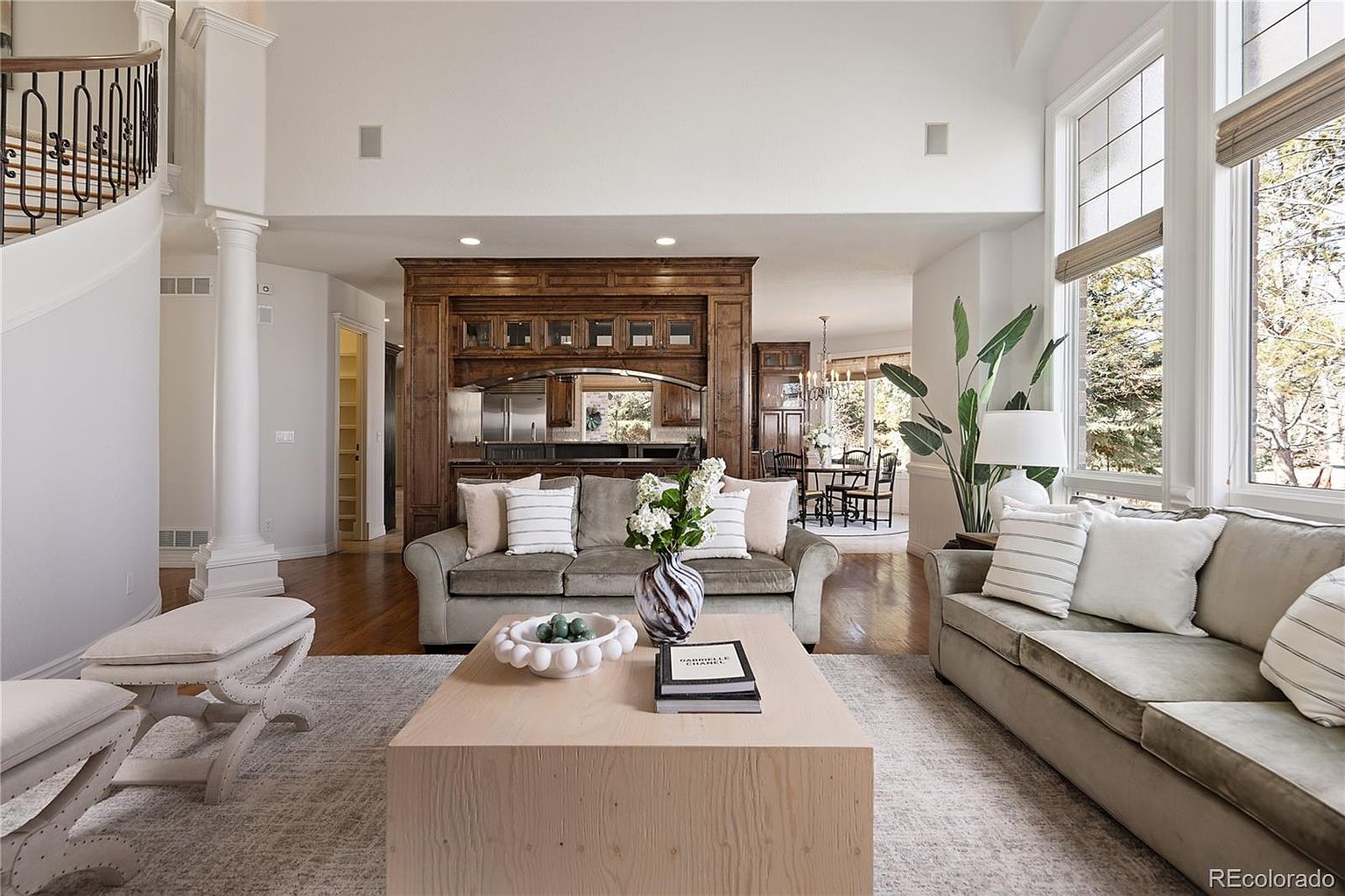 This spacious, light-filled living room features a high vaulted ceiling and an elegant curved staircase, creating an airy and sophisticated atmosphere. The room is anchored by a large, light-wood coffee table and plush, neutral-toned sofas, complemented by a rich, dark wood built-in shelving unit that adds a touch of traditional warmth. Large windows flood the space with natural light, offering a seamless transition to the dining area and highlighting the home's open-concept design.