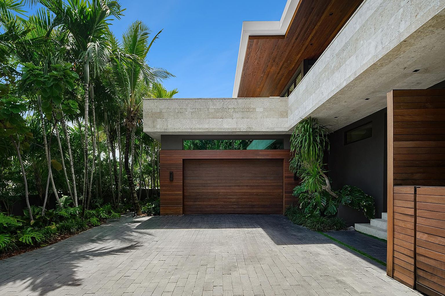 This modern residence features a prominent wood-paneled garage door set against a striking contrast of dark gray walls and light-toned stone cladding. The driveway is paved with rectangular gray stones, leading up to the garage, while lush tropical landscaping and tall palm trees frame the left side of the structure. The architectural design emphasizes clean lines and a sophisticated blend of natural textures, creating a high-end, contemporary curb appeal.