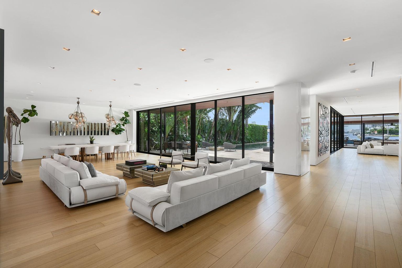 This expansive, open-concept living area features light wood flooring and a minimalist aesthetic with white walls and recessed lighting. The space is anchored by two plush, light-colored sofas facing each other around a tiered coffee table, with a long dining table visible in the background. Large, floor-to-ceiling glass sliding doors provide a seamless transition to an outdoor patio and pool area, bathing the interior in natural light.