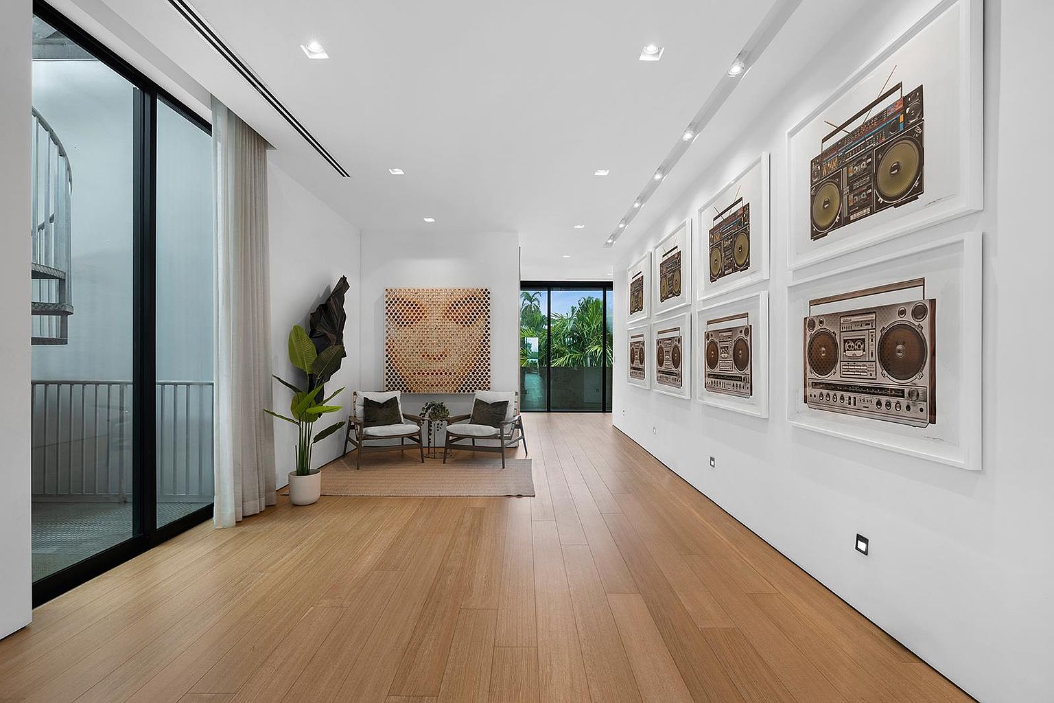 This bright and airy hallway features light wood flooring and crisp white walls, creating a clean, modern aesthetic. The space is decorated with a series of framed boombox prints on the right wall and a large, textured abstract art piece above two armchairs at the end of the hall. Large glass doors on the left provide a view of an outdoor spiral staircase, while the perspective leads the eye toward a lush green outdoor area visible through the far end of the corridor.