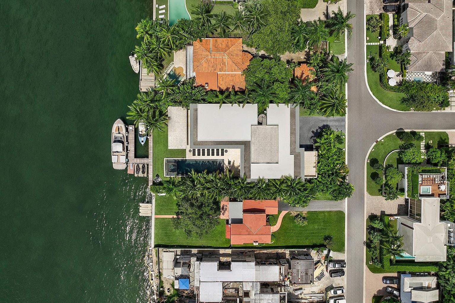 This high-altitude aerial view captures a luxurious waterfront property featuring a modern residence with a flat roof and a private swimming pool. The estate includes a private dock with a boat moored on the calm water, surrounded by lush tropical landscaping and neighboring homes. The perspective provides a comprehensive look at the property's layout, highlighting its prime location and outdoor living spaces.