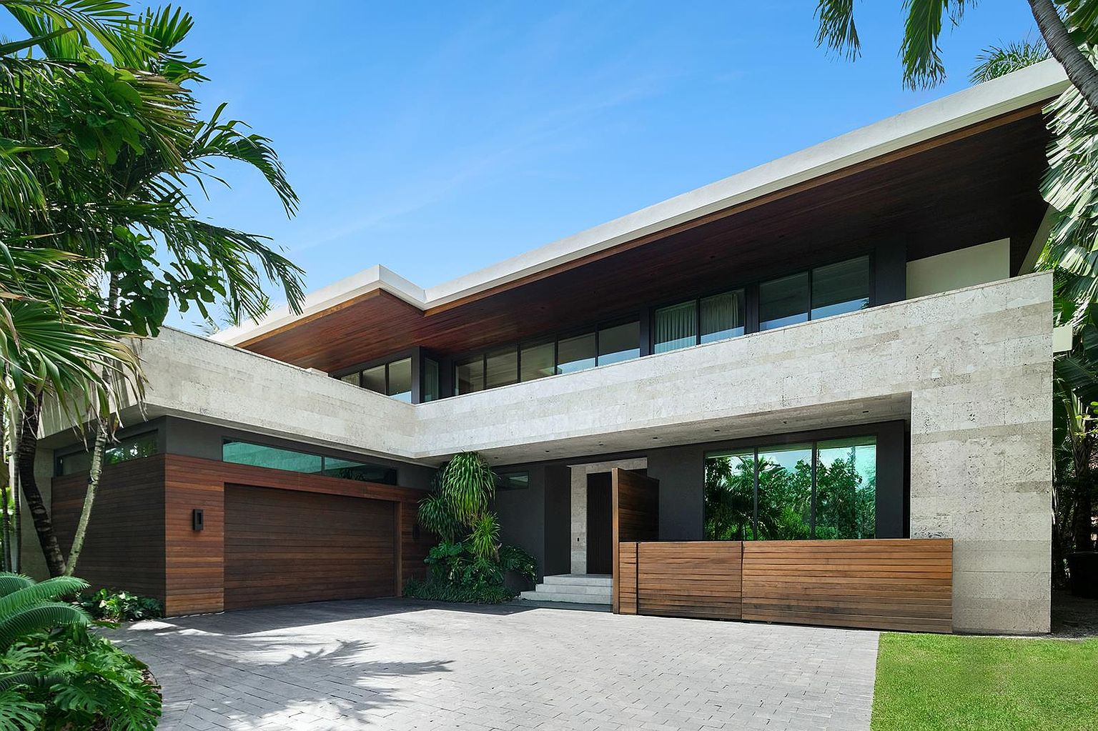 This modern residence features a striking architectural design with a combination of light-toned stone cladding and warm wood paneling. The front facade includes a large garage, a recessed entryway, and a second-story level with expansive windows under a deep, wood-lined roof overhang. Lush tropical landscaping and a paved driveway complete the sophisticated, contemporary curb appeal of this property.