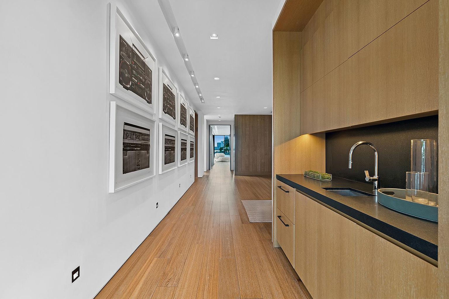 This sleek, modern hallway features light wood flooring and a minimalist aesthetic, leading the eye toward a distant doorway. To the right, a built-in wet bar area is integrated into the wall with light wood cabinetry and a dark countertop, while the left wall is adorned with a series of framed black-and-white art prints. The perspective is a straight-on shot that emphasizes the clean lines and depth of the corridor.
