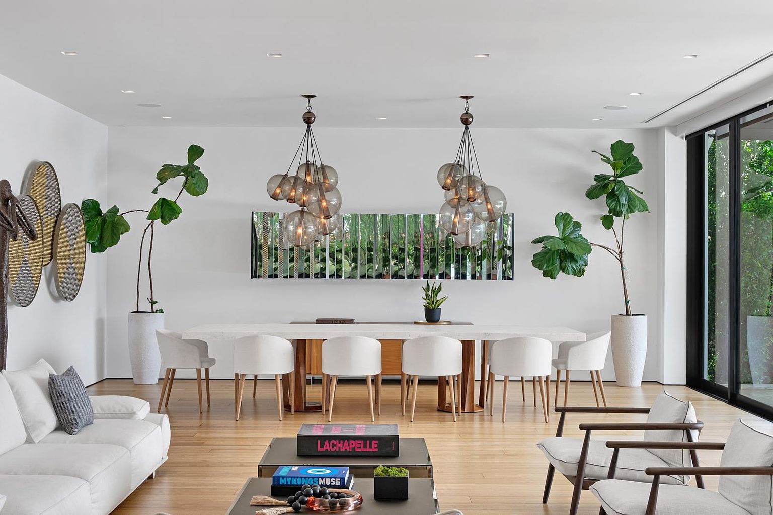 This sophisticated dining area features a long, light-toned stone dining table surrounded by six modern white chairs with light wood legs. The space is anchored by two large, multi-globe pendant light fixtures and a unique, segmented mirror wall art piece, while tall fiddle-leaf fig plants in large planters frame the room. The open-concept layout flows seamlessly into a living area, creating an airy and elegant atmosphere with warm wood flooring and large glass doors leading to the outdoors.