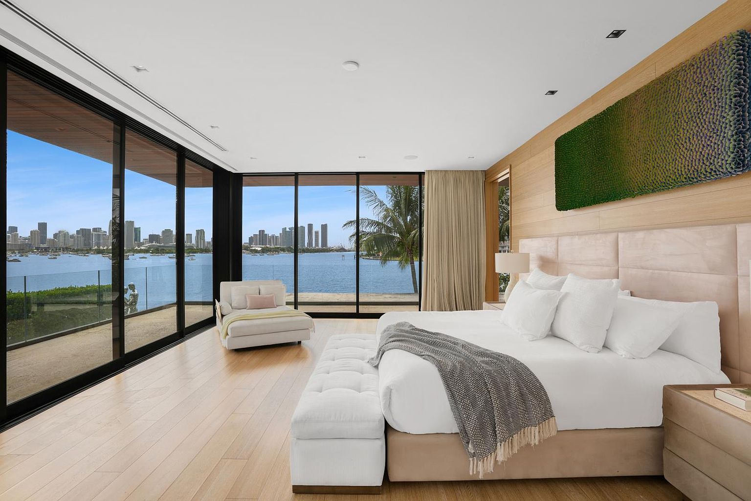 This luxurious primary bedroom features expansive floor-to-ceiling windows that offer panoramic views of the bay and city skyline. The room is anchored by a large bed with a tufted headboard, complemented by light wood flooring and a textured wall art piece. A plush lounge chair sits near the glass, creating a serene space that seamlessly blends indoor comfort with the waterfront scenery.