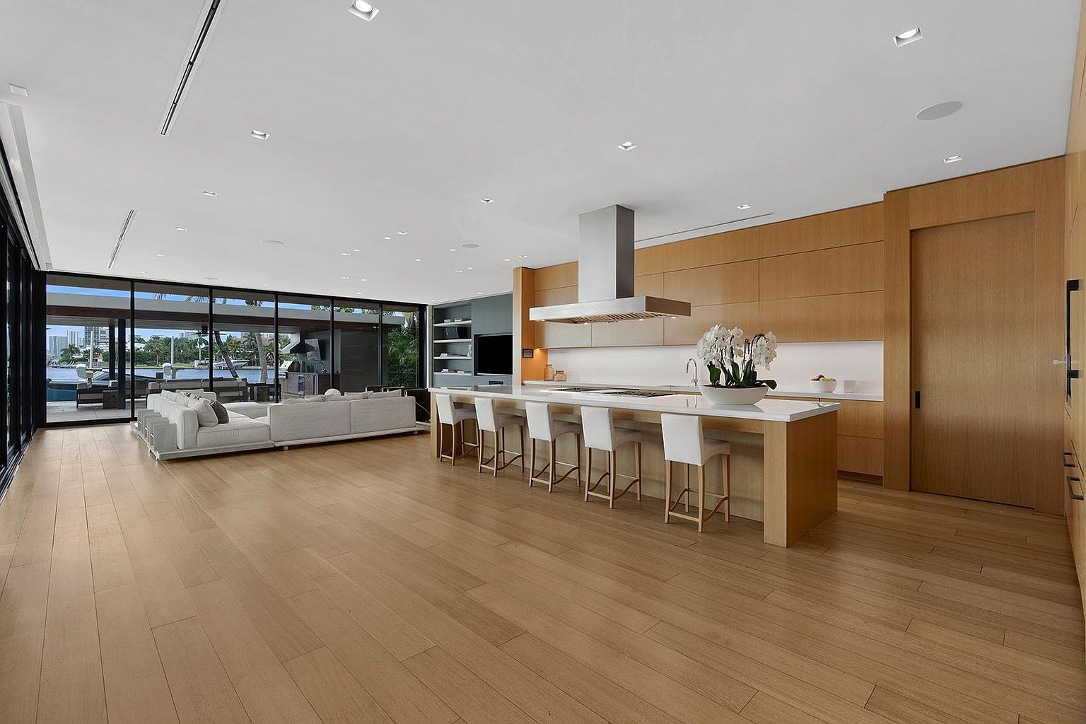 This expansive, open-concept kitchen features light wood cabinetry and a long central island with seating for five. The space is defined by its minimalist aesthetic, clean lines, and a seamless transition to the adjacent living area, which overlooks a waterfront view through floor-to-ceiling glass walls. The perspective is wide and inviting, showcasing the high-end finishes and the bright, airy atmosphere of the home.