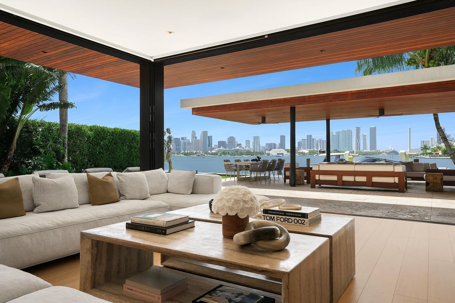 This luxurious open-concept living space seamlessly transitions to an expansive outdoor terrace, offering panoramic views of the city skyline across the water. The interior features a plush, neutral-toned sectional sofa and a substantial wooden coffee table adorned with curated books and a floral centerpiece. The architecture emphasizes indoor-outdoor living with a wide, open-air design that frames the waterfront scenery as a primary focal point.