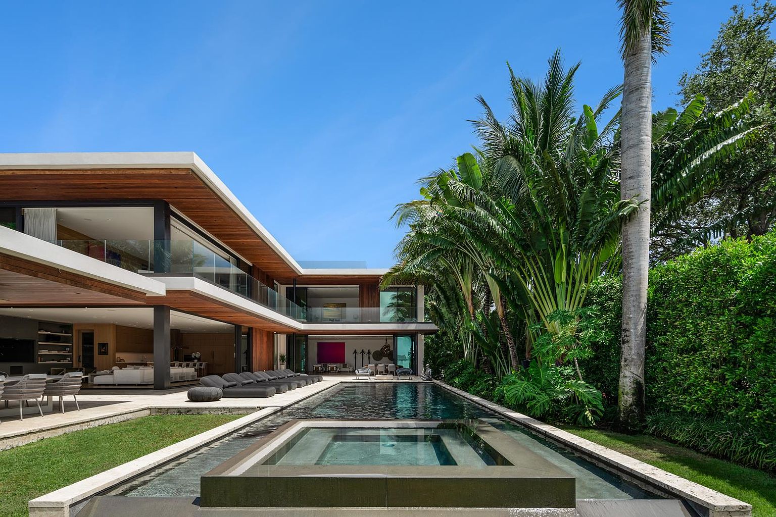 This modern luxury residence features a sleek, multi-level architectural design with extensive wood paneling and large glass walls that open to a spacious outdoor living area. A long, narrow lap pool with an integrated raised spa serves as the centerpiece of the backyard, flanked by a stone patio and lush tropical landscaping. The perspective captures the seamless transition between the indoor living space and the serene, resort-style pool deck under a clear blue sky.