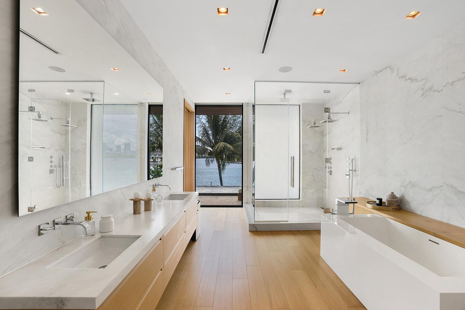 This luxurious primary bathroom features a sleek, modern design with light wood flooring and extensive stone wall cladding. The space includes a long double vanity with integrated sinks, a large glass-enclosed walk-in shower, and a deep soaking tub positioned near a window that offers a scenic water view. The perspective is centered, drawing the eye through the bright, airy room toward the outdoor scenery visible through the glass door at the far end.