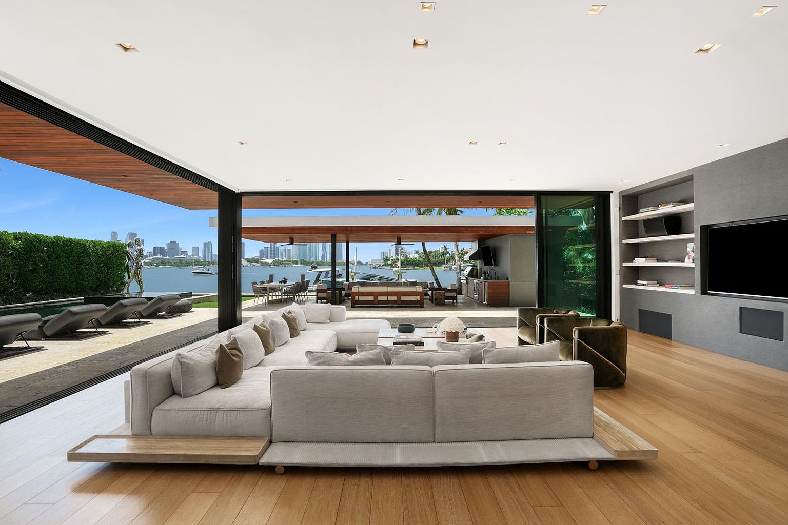 This expansive, open-concept living room features a large, low-profile sectional sofa set on a light wood floor, facing a wall with built-in shelving and a television. The space seamlessly transitions to an outdoor patio through floor-to-ceiling glass walls, offering an unobstructed view of the waterfront and city skyline. The design emphasizes a modern, indoor-outdoor lifestyle with clean lines and a neutral color palette.