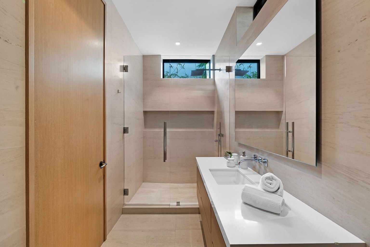 This modern bathroom features a sleek, minimalist aesthetic with light-toned stone wall cladding and matching floor tiles. The space includes a glass-enclosed walk-in shower with a recessed niche and a floating vanity topped with a white surface and a large, frameless mirror. A high, horizontal window allows natural light to filter into the room, creating a bright and serene atmosphere.
