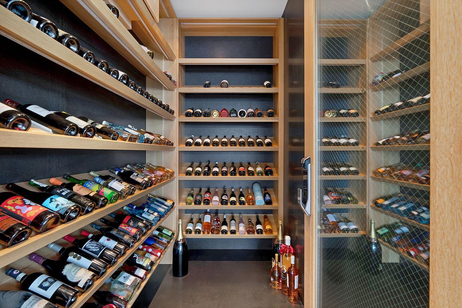 This sophisticated wine cellar features custom light wood shelving arranged in a mix of horizontal display racks and standard bottle storage. The space is divided into two sections, with an open-concept area on the left and a glass-enclosed, climate-controlled section on the right. The dark wall finish provides a dramatic backdrop that highlights the collection, creating an elegant and organized atmosphere for wine storage.