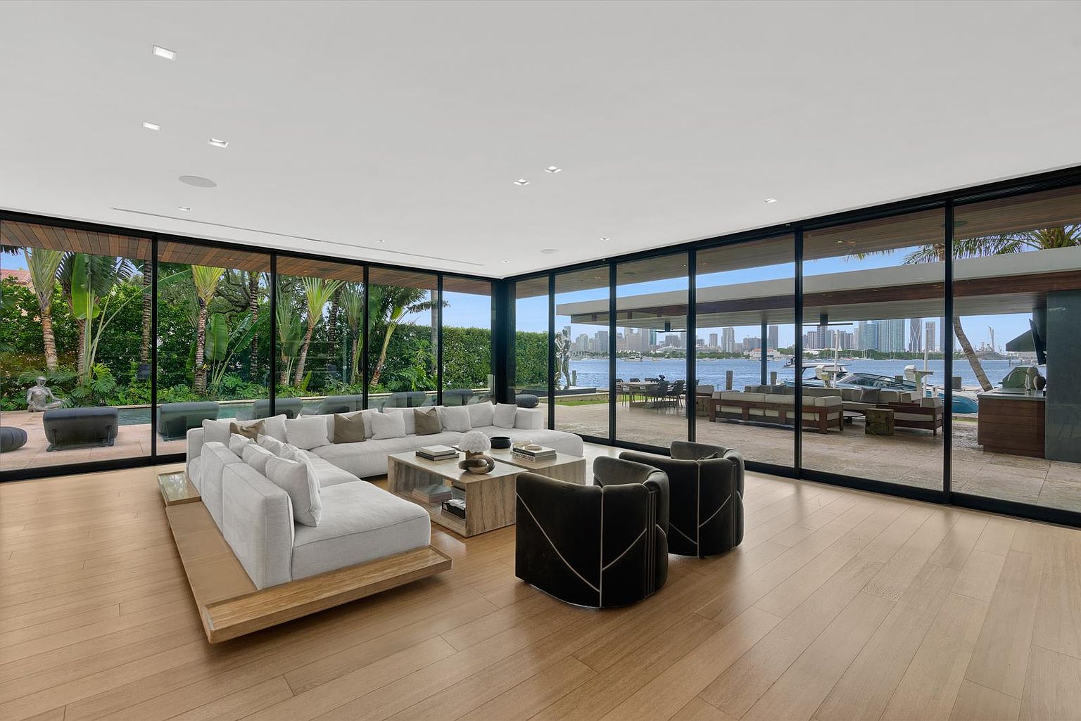 This expansive, modern living room features light wood flooring and a large, low-profile sectional sofa arranged around a central stone-topped coffee table. The space is defined by extensive floor-to-ceiling glass walls that provide panoramic views of the waterfront, pool, and lush tropical landscaping outside. Two dark, curved accent chairs complete the seating area, creating a sophisticated and airy atmosphere that seamlessly blends indoor and outdoor living.
