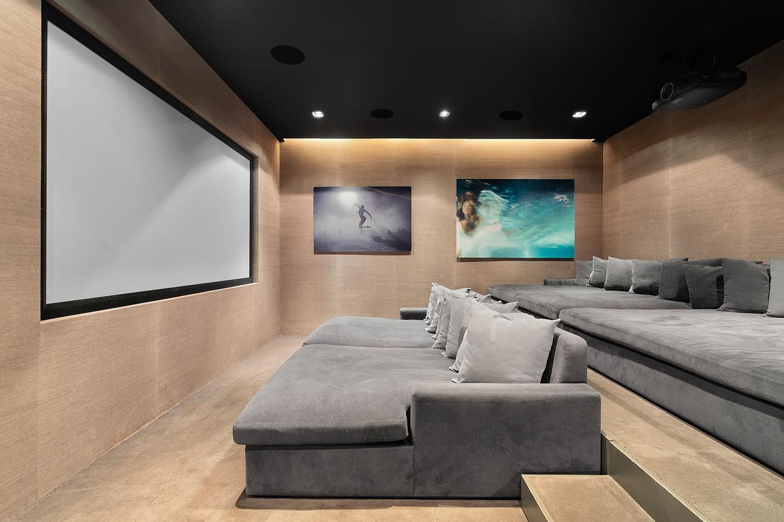 This home theater features a tiered seating arrangement with plush, grey velvet-upholstered sofas and numerous matching throw pillows. The walls are finished in a warm, light-toned wood paneling, complemented by a dark ceiling and recessed lighting. A large projection screen dominates the front wall, while two framed art pieces add a decorative touch to the side wall.