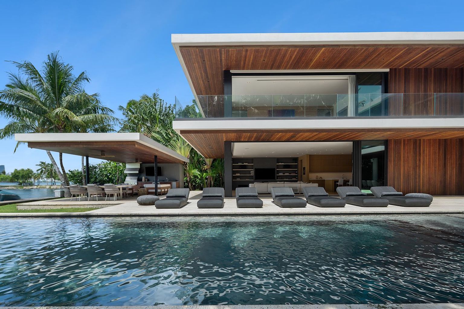 This stunning modern residence features a seamless indoor-outdoor transition, highlighted by a large swimming pool that reflects the architectural lines of the home. The exterior showcases extensive use of warm-toned wood siding and deep, cantilevered roof overhangs that provide shade to the poolside lounge area. A separate covered outdoor kitchen and dining space sits adjacent to the main structure, creating an ideal environment for luxury waterfront entertaining.