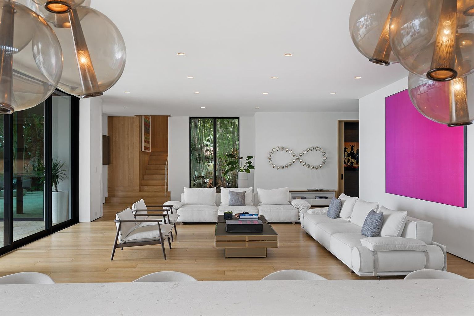 This spacious, contemporary living room features light wood flooring and a bright, open-concept layout. The space is anchored by two large white sofas and a pair of modern armchairs, centered around a low-profile coffee table, with a striking infinity-shaped wall sculpture serving as a focal point. Large glass sliding doors provide a seamless connection to the outdoors, while a bold magenta art piece adds a vibrant splash of color to the minimalist white walls.