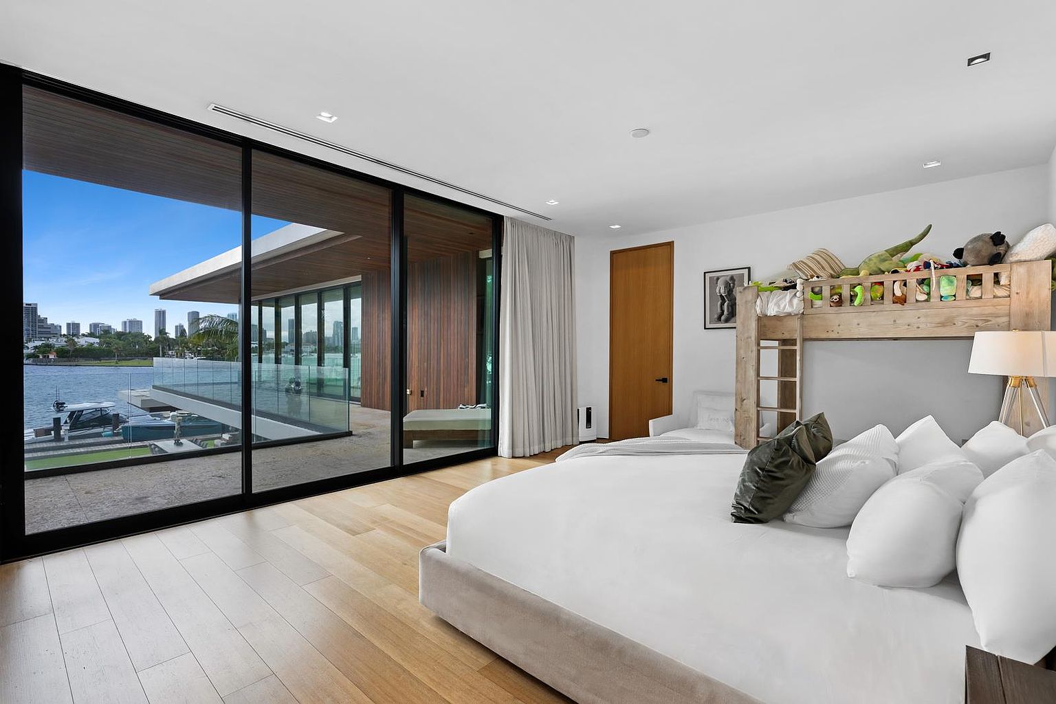 This modern bedroom features light wood flooring and a large bed paired with a wooden bunk bed structure adorned with stuffed animals. Expansive floor-to-ceiling glass sliding doors provide a seamless transition to a spacious waterfront balcony with views of the bay and city skyline. The room is bright and airy, characterized by a minimalist aesthetic and neutral color palette.