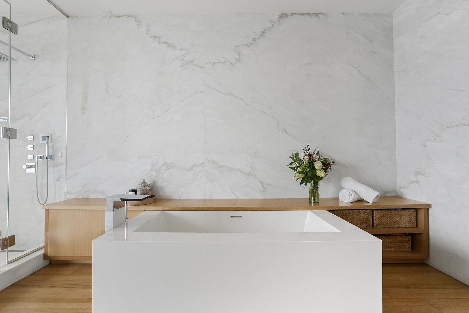 This primary bathroom features a minimalist, spa-like aesthetic centered around a large, white rectangular soaking tub. The space is anchored by a wall clad in large-format stone panels with subtle veining, complemented by a light wood vanity surface and integrated shelving. To the left, a glass-enclosed shower area adds to the open, airy feel, while a vase of fresh flowers provides a touch of organic warmth to the clean, modern design.