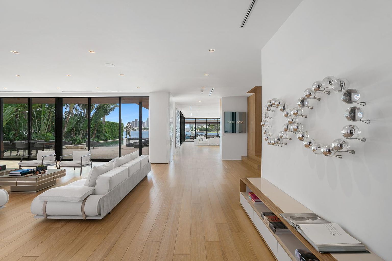 This expansive, open-concept living area features light wood flooring and a minimalist aesthetic, anchored by a large white sectional sofa. A striking, abstract metallic wall sculpture serves as a focal point, while floor-to-ceiling glass walls provide a seamless transition to the outdoor pool and waterfront views. The perspective looks through the bright, airy space toward a secondary living area, emphasizing the home's generous scale and modern architectural design.