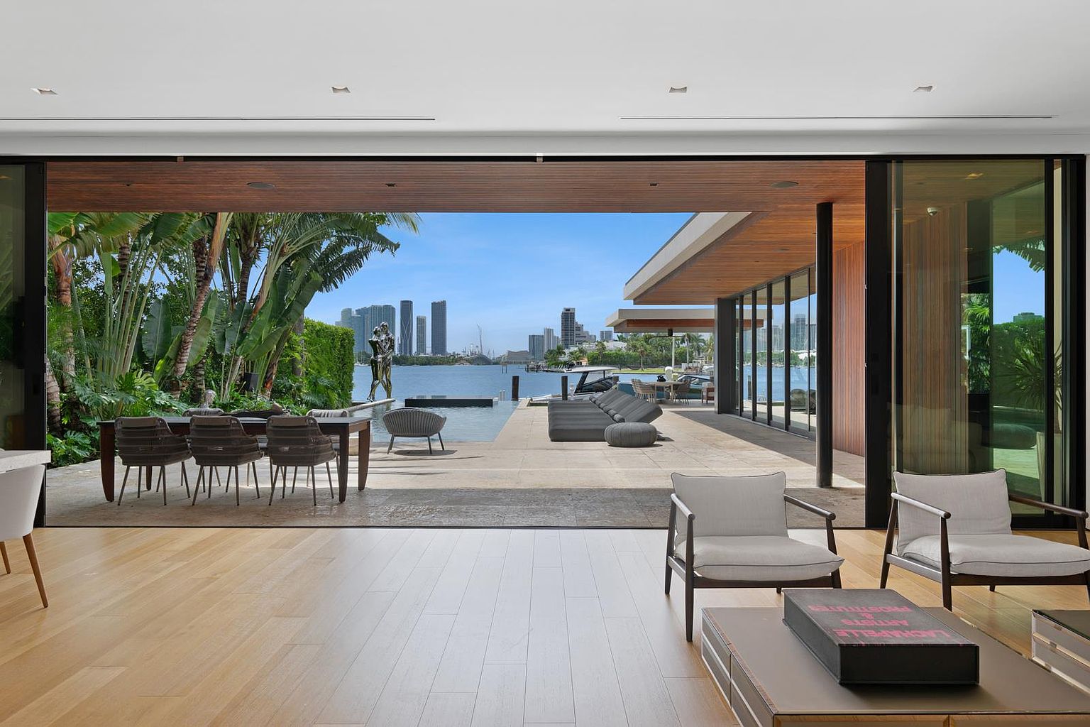 This expansive living area features light wood flooring and seamless indoor-outdoor integration through massive sliding glass walls that open onto a waterfront patio. The space is furnished with modern lounge chairs and a low-profile coffee table, while the exterior deck showcases a dining set, a large sectional, and a sculpture overlooking the bay and city skyline. The overall aesthetic is one of sophisticated, contemporary luxury designed for effortless entertaining.