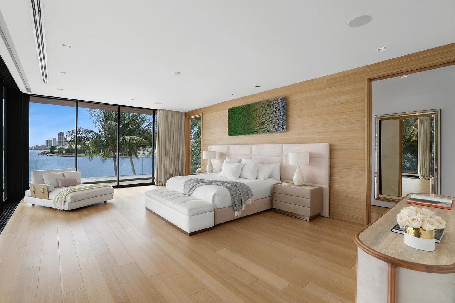 This expansive primary bedroom features light wood flooring and a wall of floor-to-ceiling glass that frames a serene waterfront view. The room is anchored by a large bed with a neutral upholstered headboard, complemented by a matching bench and a contemporary chaise lounge positioned near the window. A textured green art piece hangs above the bed, while a sleek console table with a floral arrangement sits in the foreground, creating a sophisticated and tranquil atmosphere.