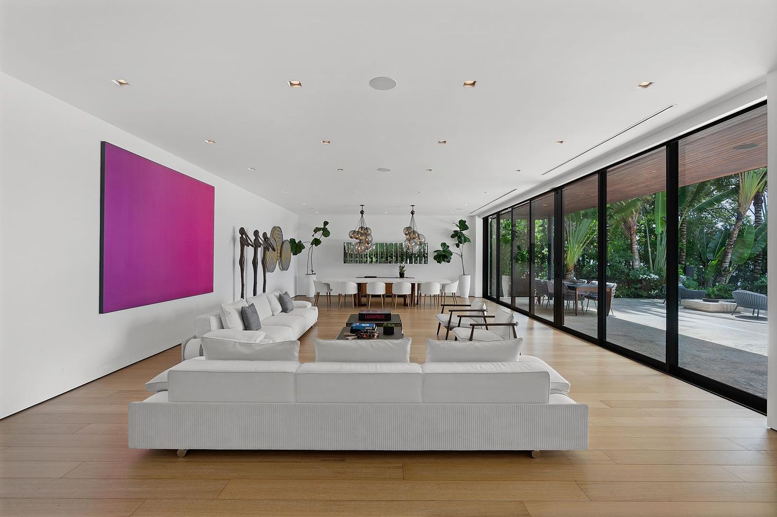 This expansive, open-concept living and dining area features light wood flooring and a minimalist aesthetic. A large, vibrant purple art piece serves as a focal point on the white wall, while floor-to-ceiling glass walls provide a seamless transition to the lush outdoor greenery. The space is furnished with plush white sofas and a sleek dining set, illuminated by modern pendant lighting and recessed ceiling lights.