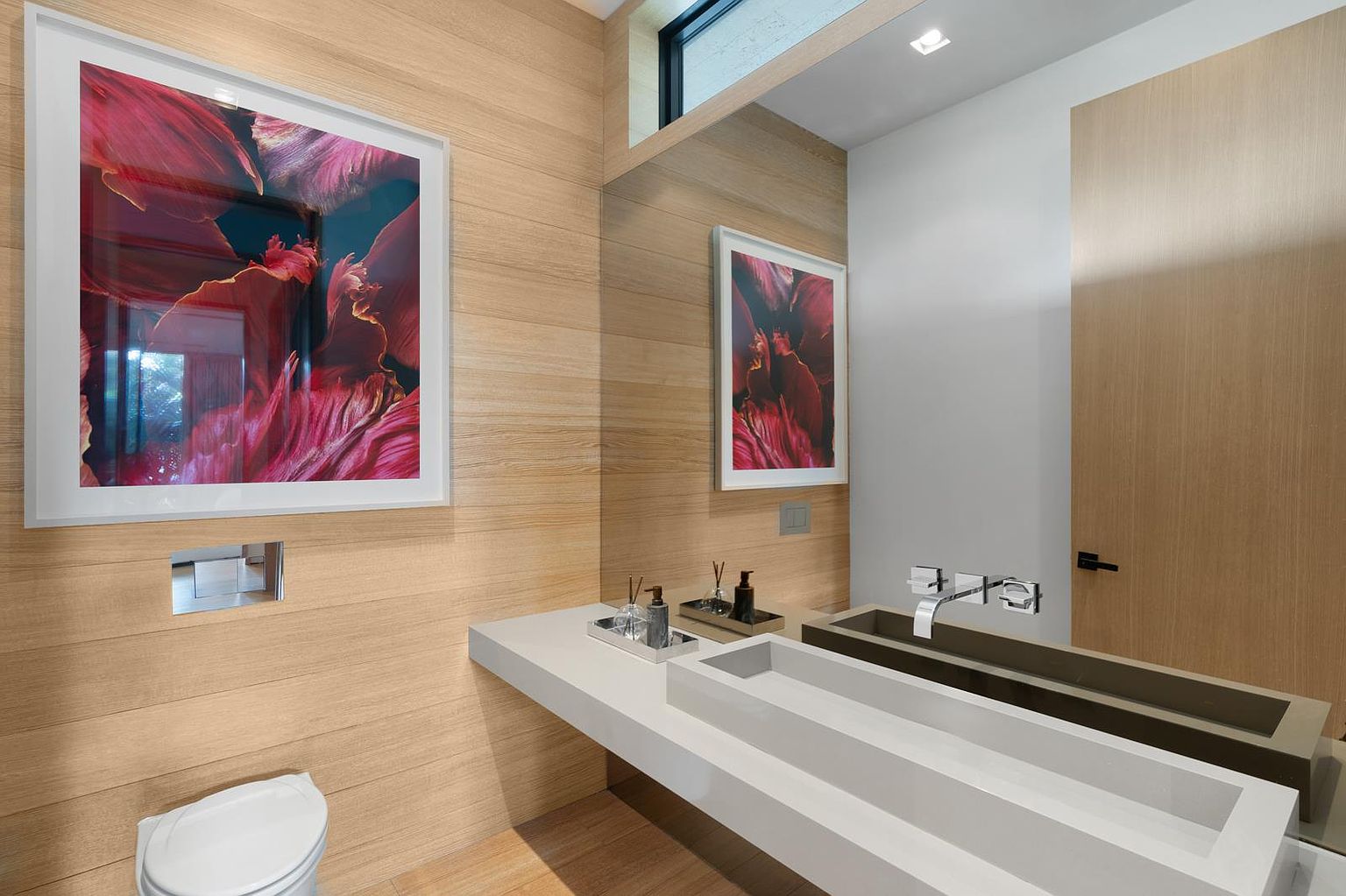 This modern powder room features light wood-paneled walls and a minimalist white trough-style sink mounted on a floating vanity. A large, vibrant floral art piece is displayed on the wall, reflected in the expansive mirror that spans the vanity area. The space is accented by sleek chrome fixtures and a wall-mounted toilet, creating a clean, contemporary aesthetic.