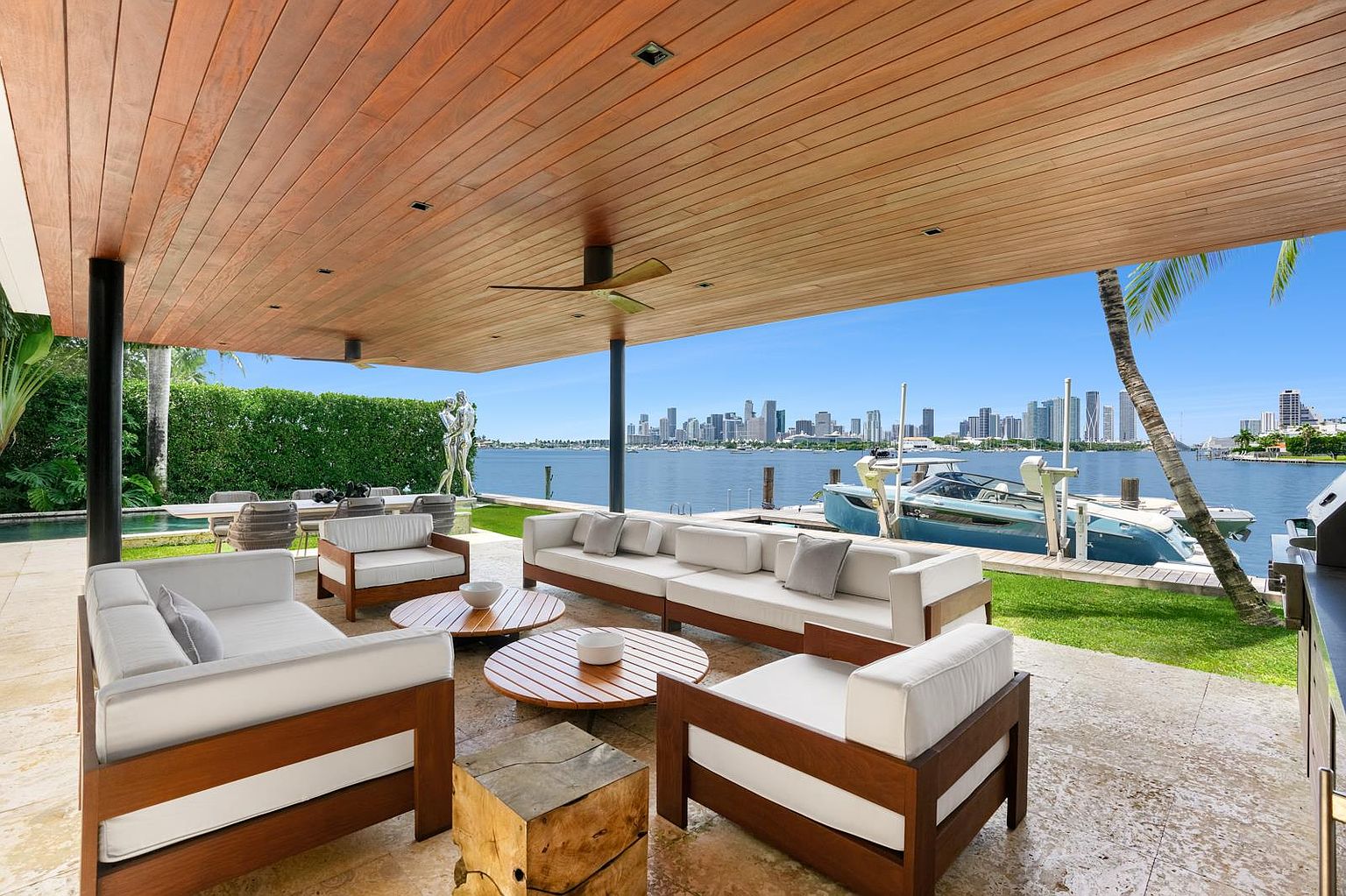 This expansive, covered outdoor living space features a warm wood-paneled ceiling and light-toned stone flooring, creating a seamless transition to the waterfront. The area is furnished with modern, low-profile seating arrangements and circular wooden coffee tables, perfectly positioned to enjoy the panoramic city skyline view across the water. A private boat dock with a docked vessel sits just beyond the manicured lawn, emphasizing the luxury coastal lifestyle.