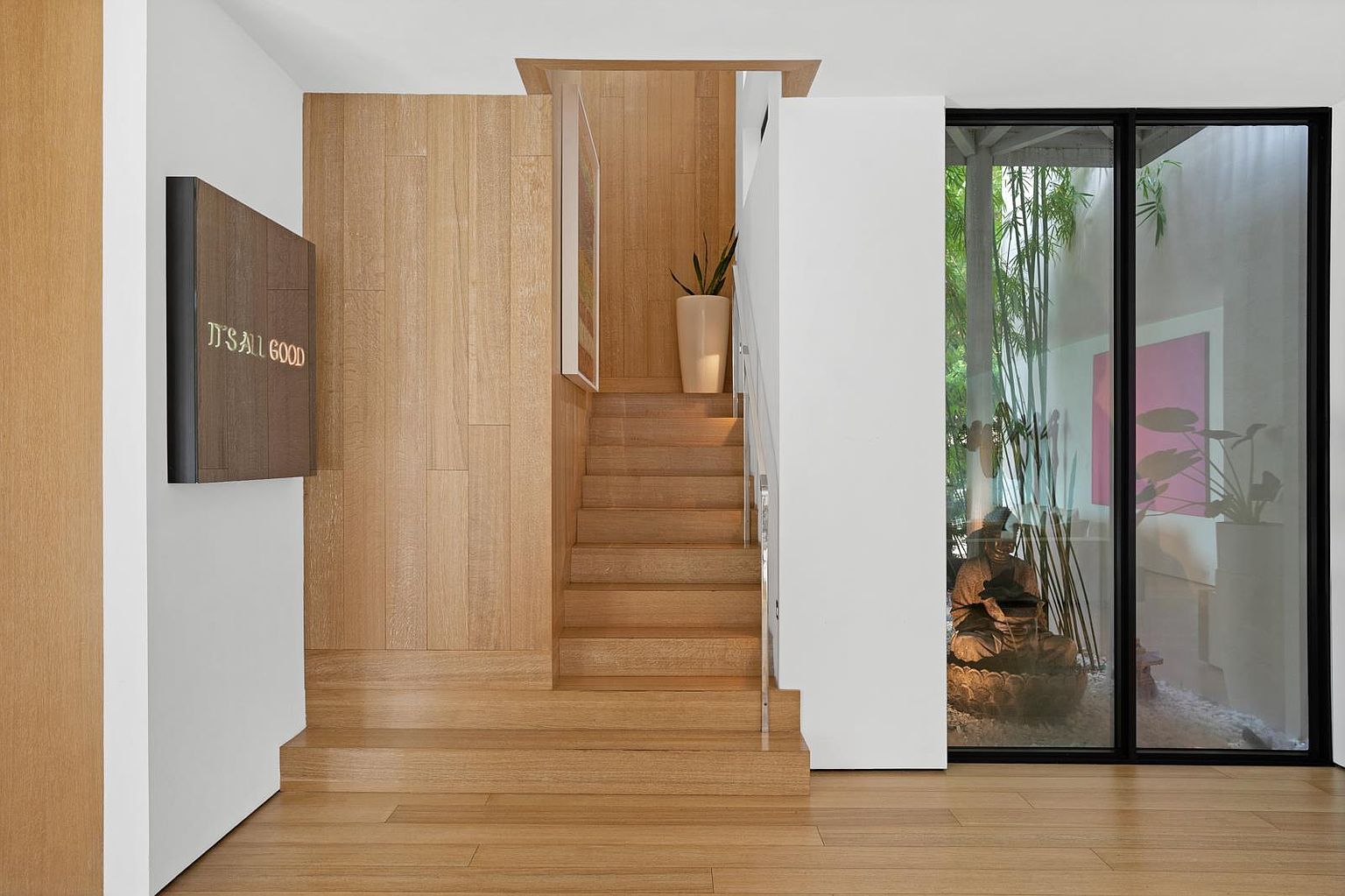 This interior view showcases a minimalist staircase featuring light wood paneling that extends up the wall, creating a warm and cohesive aesthetic. To the right, a large glass panel provides a view into an enclosed courtyard with bamboo and a decorative statue, while a sign on the left wall adds a modern touch. The perspective is centered and symmetrical, emphasizing the clean lines and natural textures of the entryway.