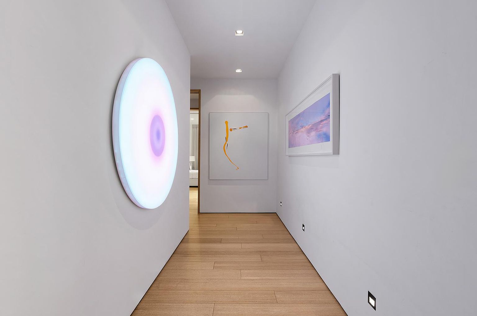 This modern, minimalist hallway features light wood flooring and crisp white walls, creating a bright and gallery-like atmosphere. The space is adorned with contemporary art, including a large, circular illuminated wall piece on the left and a framed landscape print on the right. Recessed ceiling lighting and low-profile wall-mounted lights provide a clean, sophisticated aesthetic that leads the eye toward an open doorway at the end of the hall.