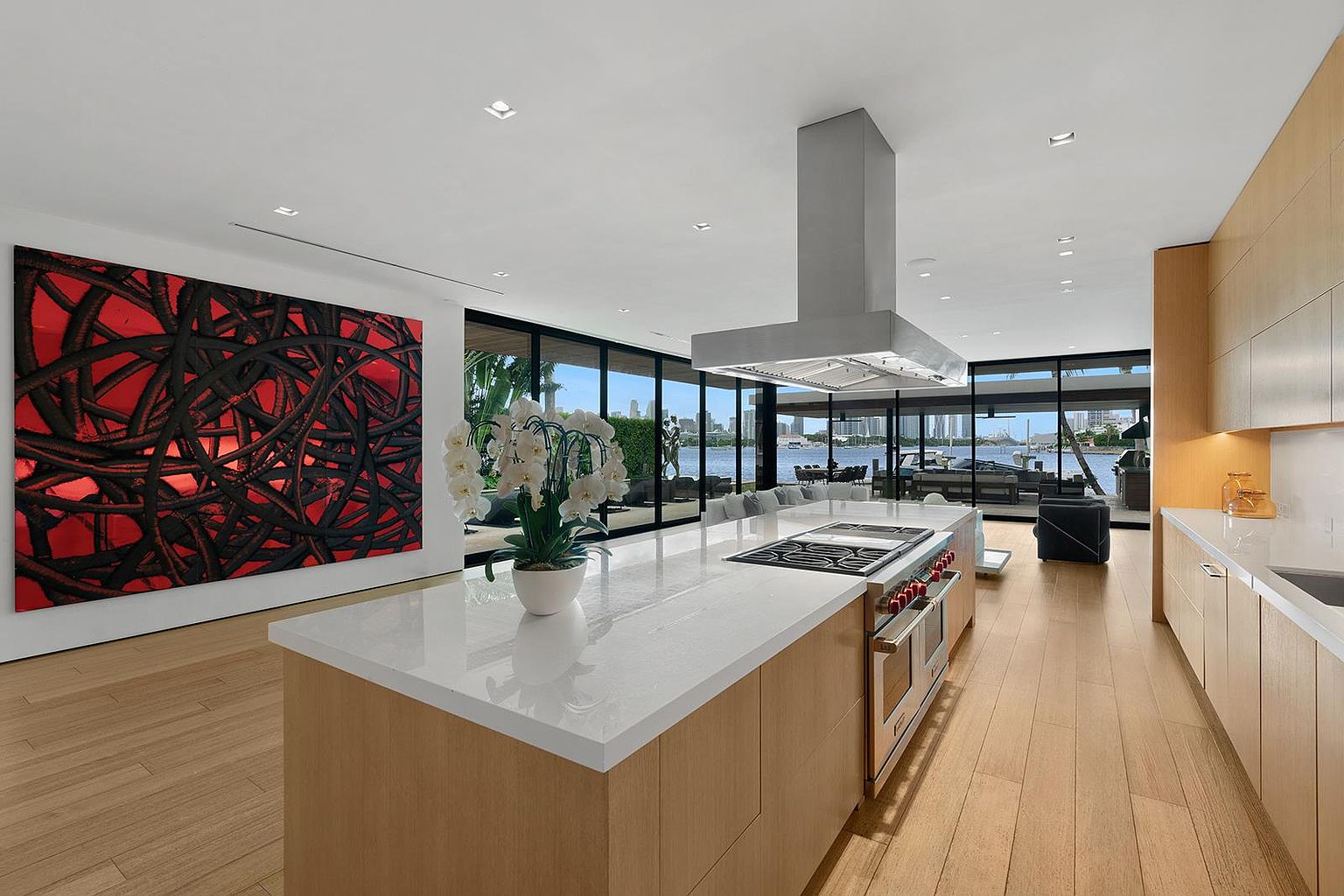This modern, open-concept kitchen features light wood cabinetry and a large central island topped with a white stone surface. A professional-grade range is integrated into the island, complemented by a stainless steel range hood suspended from the ceiling. The space is framed by expansive floor-to-ceiling windows that provide a panoramic view of the waterfront, while a large, bold abstract painting adds a striking focal point to the room.