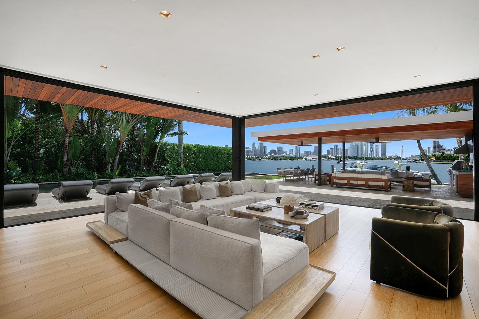 This expansive, open-concept living room features a seamless transition to the outdoors through large, retractable glass walls that frame a stunning waterfront view of the city skyline. The space is anchored by a massive, light-toned sectional sofa and low-profile wooden coffee tables, creating a relaxed, modern atmosphere. The interior flows directly onto a covered patio and pool deck, emphasizing a sophisticated indoor-outdoor lifestyle perfect for entertaining.