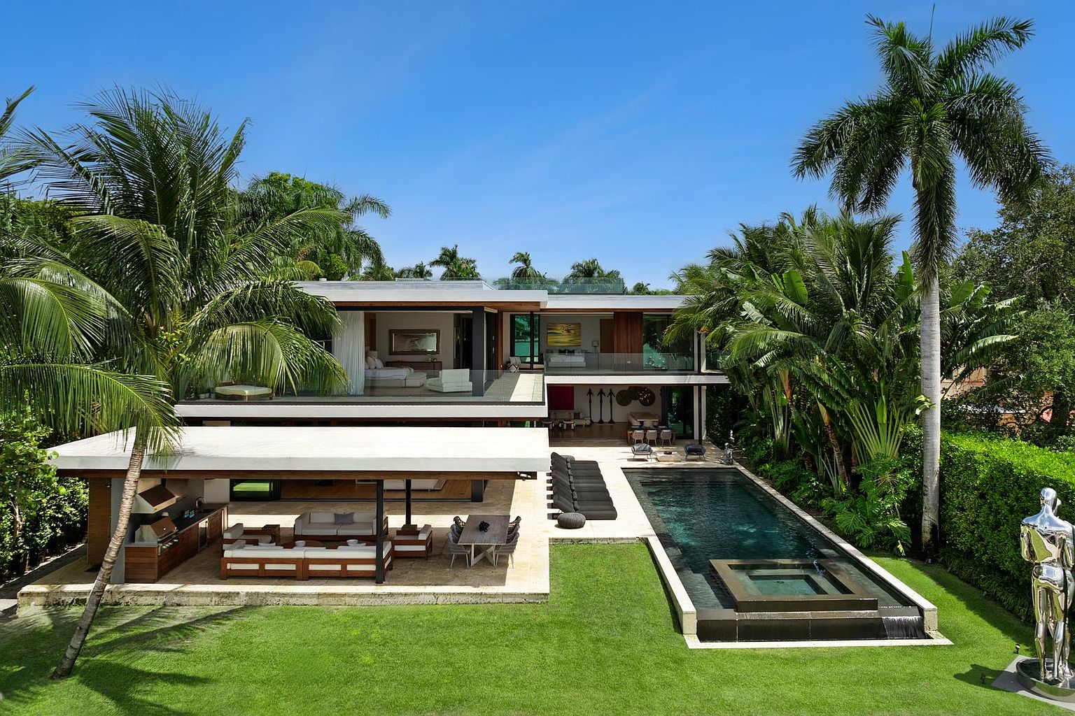 This modern luxury residence features a multi-level architectural design with expansive glass walls and deep, covered outdoor living spaces. The rear yard includes a sleek, dark-tiled rectangular pool with an integrated spa, a manicured lawn, and a covered patio area equipped with an outdoor kitchen and dining space. Lush tropical landscaping, including tall palm trees, frames the property, creating a private and sophisticated resort-like atmosphere.