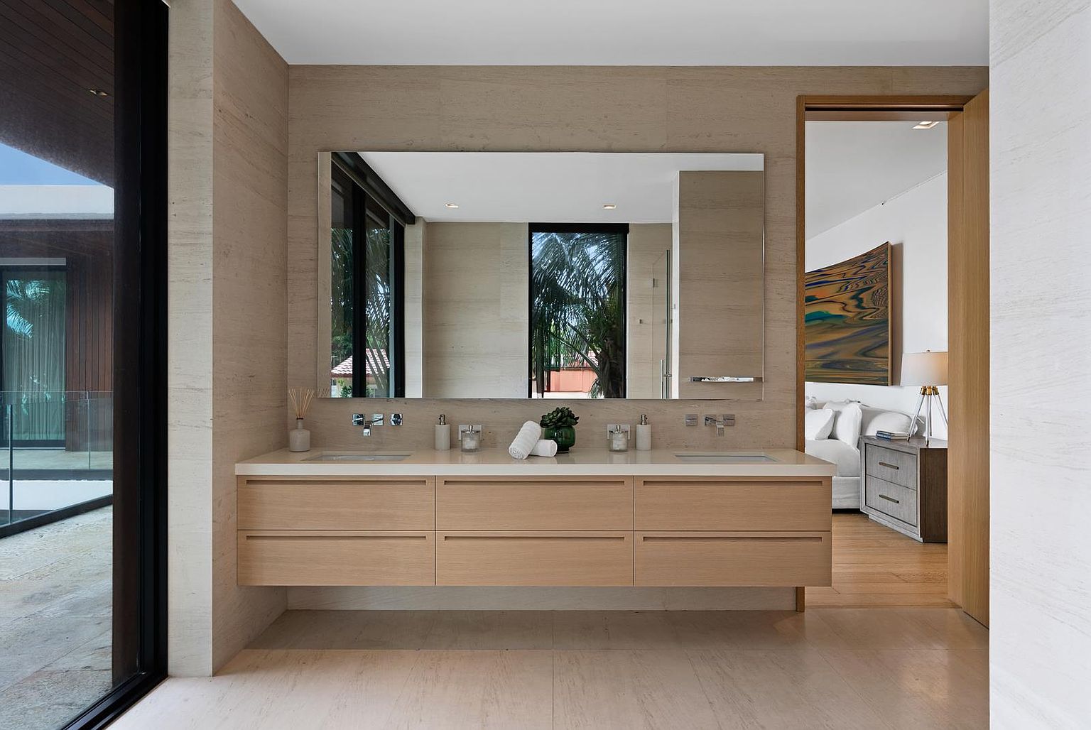 This primary bathroom features a floating double vanity with light wood cabinetry and a clean, minimalist aesthetic. A large, rectangular mirror spans the width of the vanity, reflecting the adjacent bedroom and outdoor greenery visible through a window. The space is finished with neutral stone wall tiles and flooring, creating a serene and cohesive atmosphere.