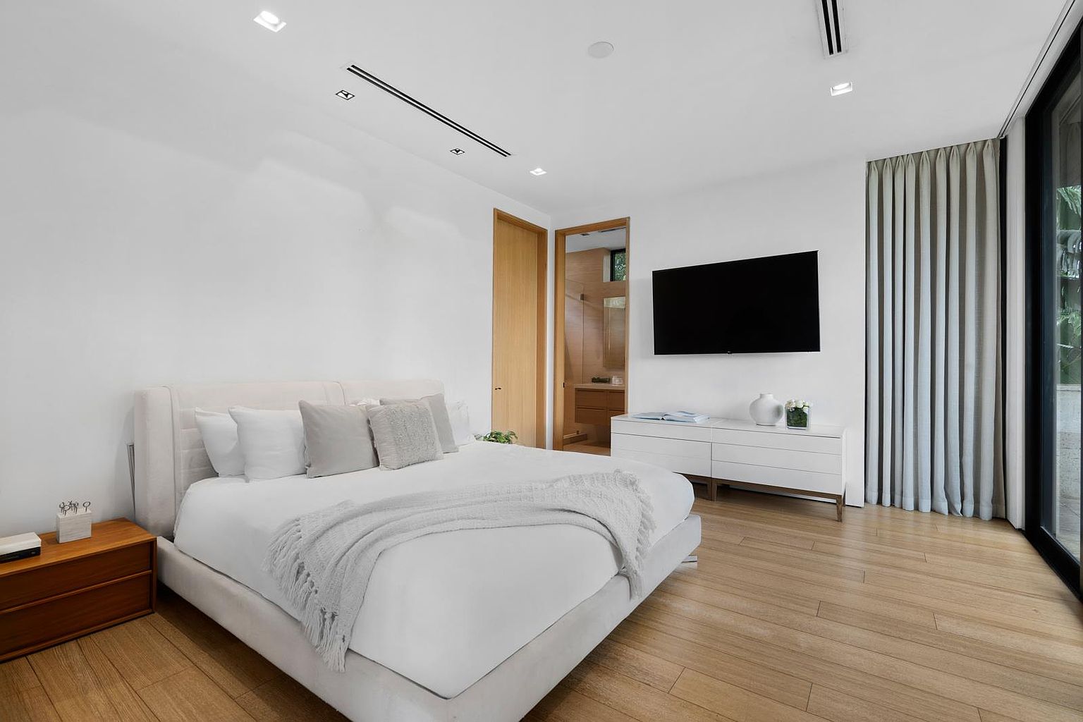 This bright and airy primary bedroom features light wood flooring and a neutral color palette, centered around a plush upholstered bed with layered linens. A sleek white dresser sits beneath a wall-mounted television, while floor-to-ceiling curtains frame a large glass door leading to an outdoor space. The room is accented by warm wood-toned doors and minimalist recessed lighting, creating a serene and contemporary atmosphere.