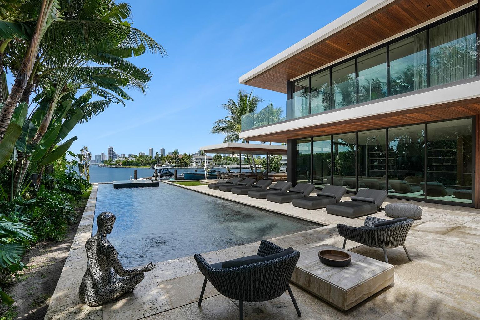 This luxurious waterfront property features a sleek, modern infinity-edge pool that seamlessly blends with the bay view in the background. The expansive stone-paved patio is furnished with multiple lounge chairs and contemporary seating, creating an ideal space for outdoor relaxation and entertaining. The architecture showcases clean lines, extensive glass walls, and warm wood accents, all set against a backdrop of lush tropical landscaping and a distant city skyline.