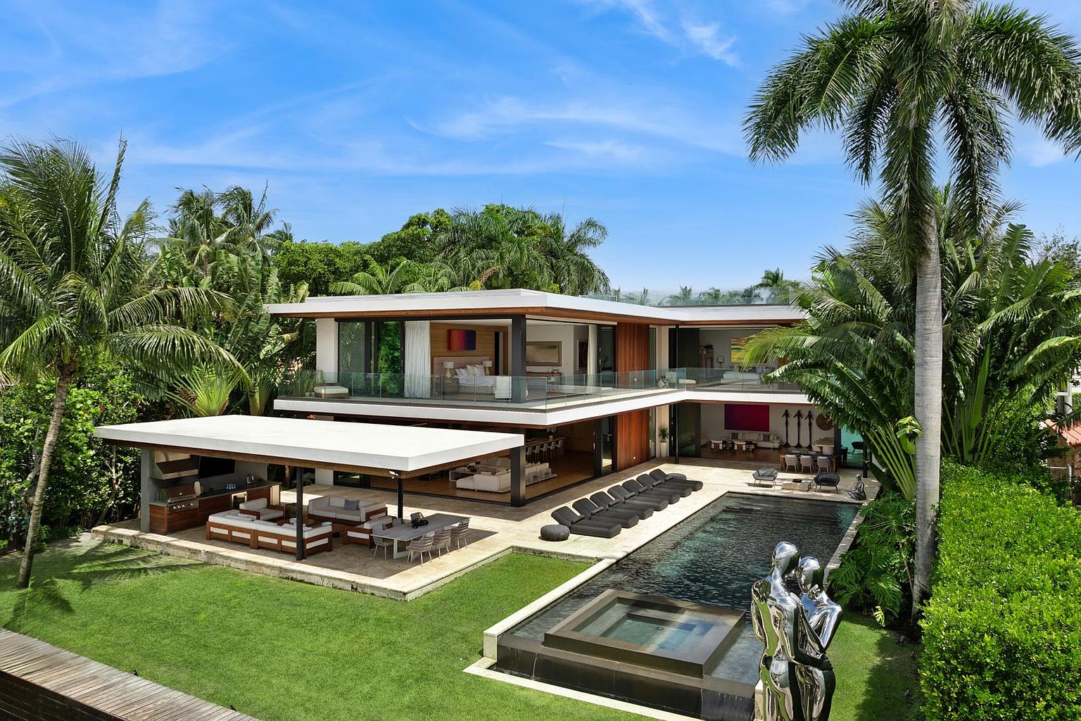 This high-angle aerial view captures a luxurious modern residence characterized by clean lines, expansive glass walls, and a seamless indoor-outdoor flow. The property features a large covered outdoor living area with a kitchen, a sleek swimming pool with an integrated spa, and a lush, tropical landscaped yard. The architecture emphasizes open-concept living, with balconies on the upper level overlooking the pool deck and manicured lawn.