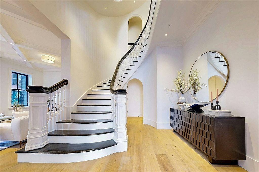 This interior shot showcases a grand foyer with a curved staircase featuring dark treads and a white banister. A dark wood console table with decorative elements sits against the wall, complemented by a round mirror and floral arrangement. The hardwood flooring and neutral wall colors create a bright and elegant atmosphere.