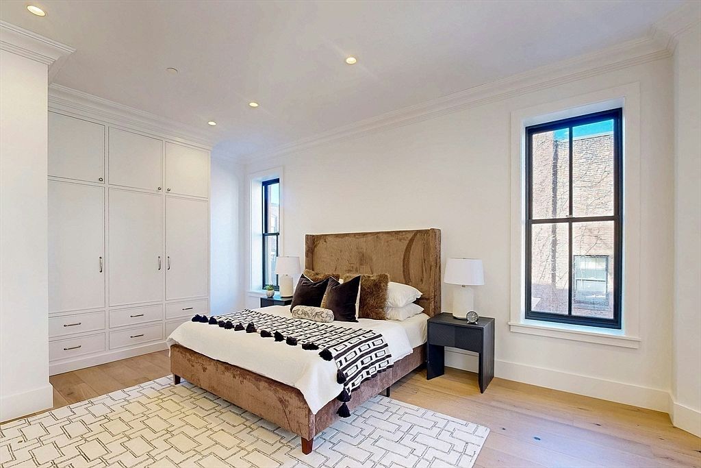 This is a well-lit primary bedroom featuring a brown upholstered bed with decorative pillows and a patterned throw. The room has built-in white wardrobes, hardwood floors, and a neutral-toned rug. Natural light streams in through two windows, complementing the recessed lighting.