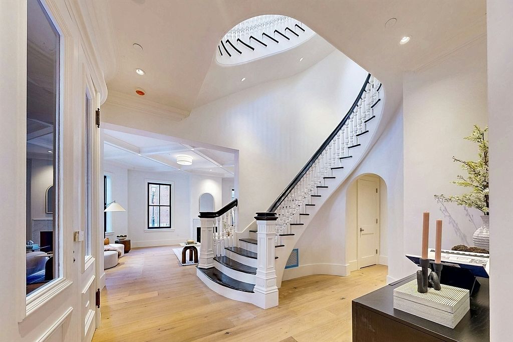 This is an interior shot of a grand hallway featuring a curved staircase with black handrails and white balusters. The walls are painted in a clean white, and the hardwood floors add warmth to the space. A unique circular skylight provides natural light, and the open layout leads into a living area, creating an inviting and luxurious atmosphere.