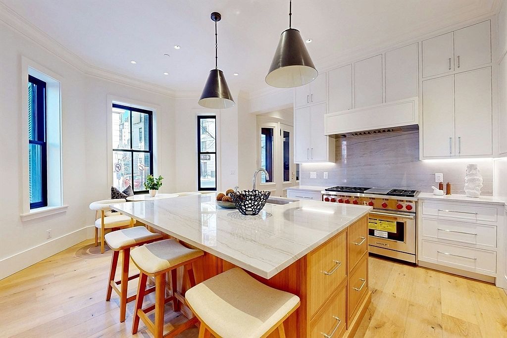 This is a bright and modern kitchen featuring a large island with a marble countertop and wooden base, providing ample seating. The kitchen is equipped with white cabinetry, stainless steel appliances including a Wolf range, and pendant lighting. Natural light floods the space through several windows, enhancing the clean and contemporary aesthetic.