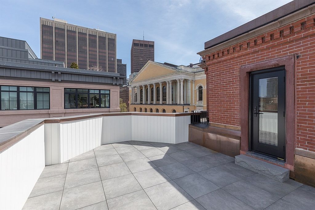 This image showcases a spacious rooftop patio with gray tiled flooring and white paneled walls. A brick building with a black door is visible on the right, while other buildings can be seen in the background, creating an urban setting. The patio offers an outdoor living space with a view of the city skyline.