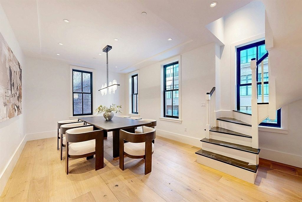 This is an interior shot of a dining room featuring a dark, square dining table with six chairs. The room has light hardwood floors, white walls, and black-framed windows that provide natural light. A modern chandelier hangs above the table, and a staircase is visible in the background, adding architectural interest.