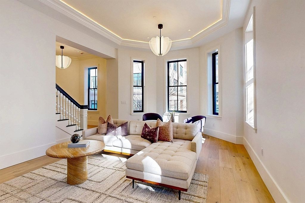 This is an interior shot of a living room featuring a sectional sofa with tufted cushions and decorative pillows, a round coffee table with books and a decorative object, and a patterned area rug. The room has hardwood floors, white walls, and large windows with black frames, providing ample natural light. A modern chandelier hangs from the ceiling, and a staircase is visible in the background.